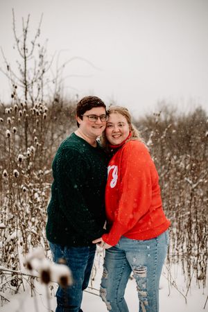 Smiling couple posing together in a snowy field, wearing coordinating red and green holiday sweaters.