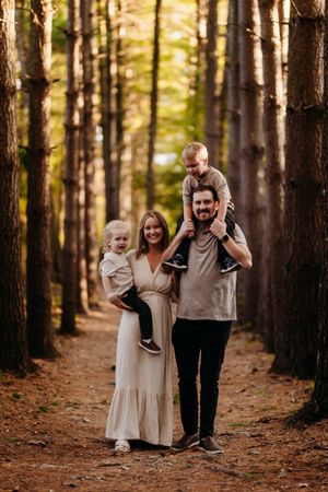 Smiling family of four in pine forest, father with son on shoulders, mother holding toddler.