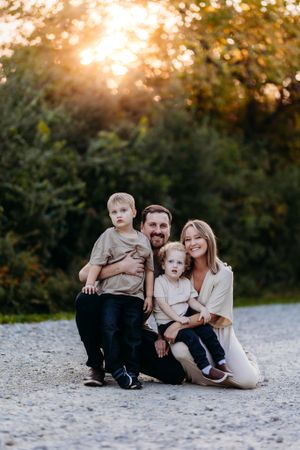 Happy family of four sitting together on gravel path with golden sunset glowing through trees.