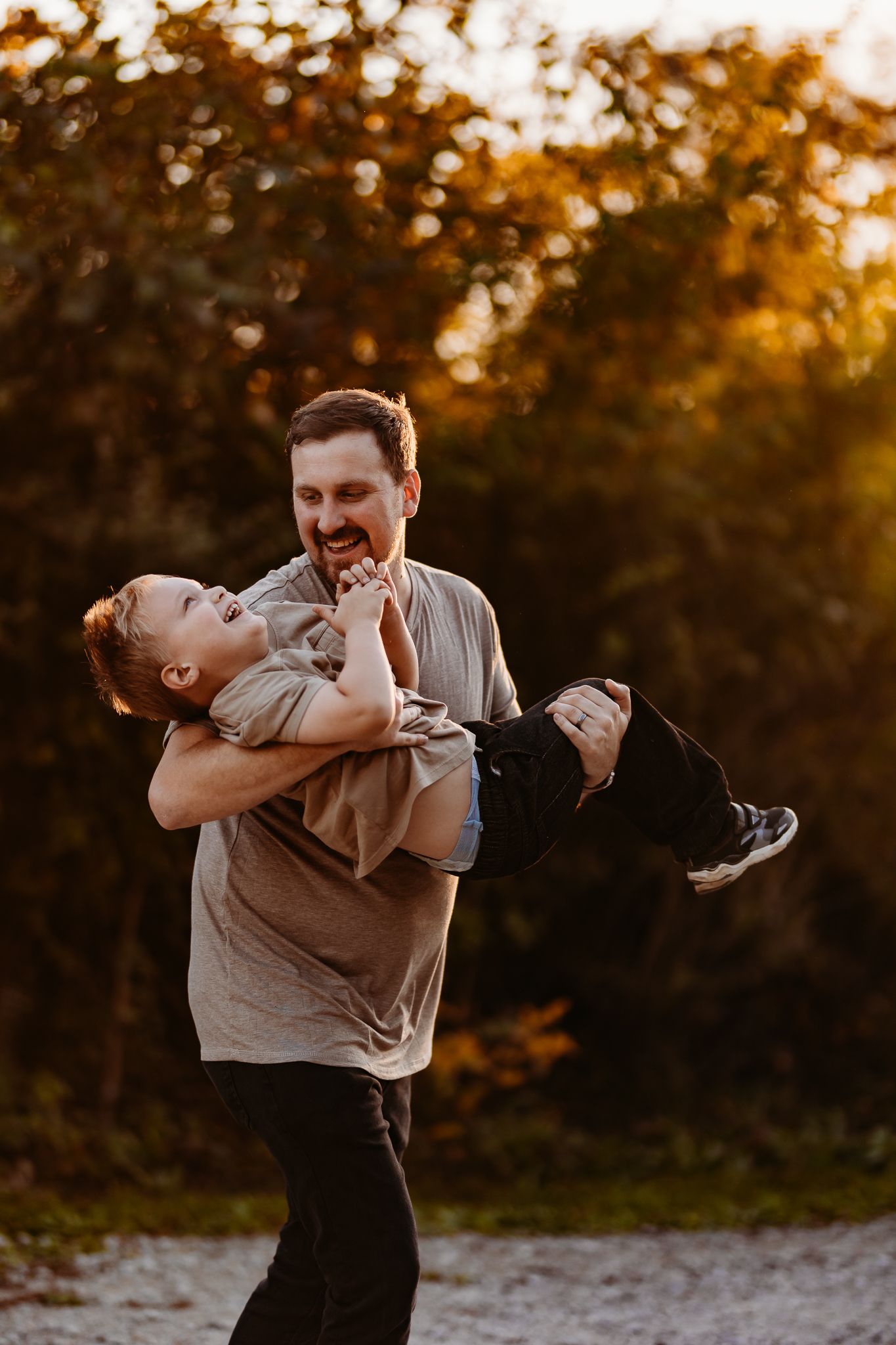 A laughing father playfully holds his giggling toddler son outdoors during a warm golden sunset.