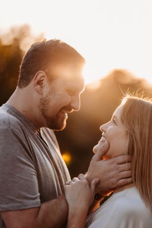 A bearded man tenderly holds a smiling womans face during a romantic golden sunset outdoors.