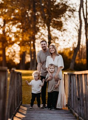 A happy family of four poses together on a wooden bridge surrounded by autumn trees at sunset.