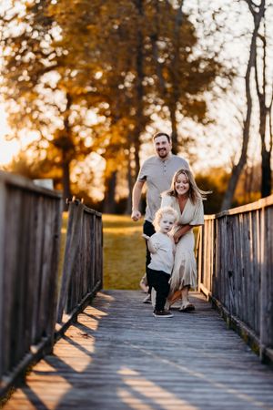 A joyful family walks along a wooden bridge at golden hour with warm autumn trees in background.