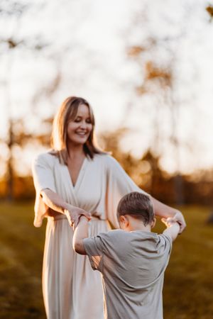 A smiling mother in a white dress holds hands with her young son in a sunlit autumn park.