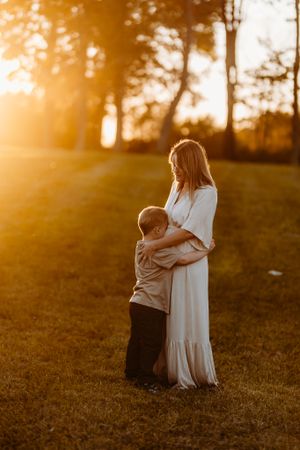 A young boy tenderly hugs his mother in a white dress during a glowing golden sunset outdoors.