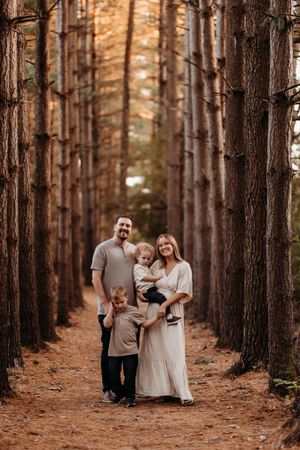 A smiling family of four poses together on a pine forest path in warm golden autumn light.