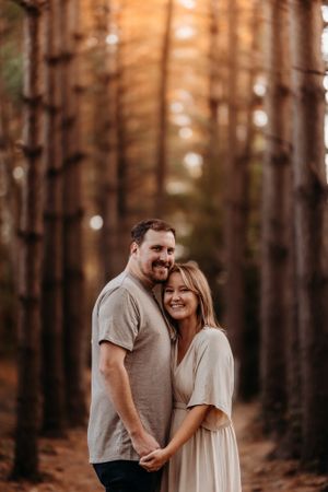 Couple embracing and smiling in a sunlit pine forest during golden hour.