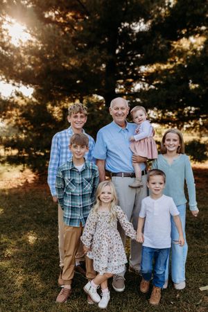 Grandfather proudly posing with six grandchildren of various ages in a sunny outdoor setting.