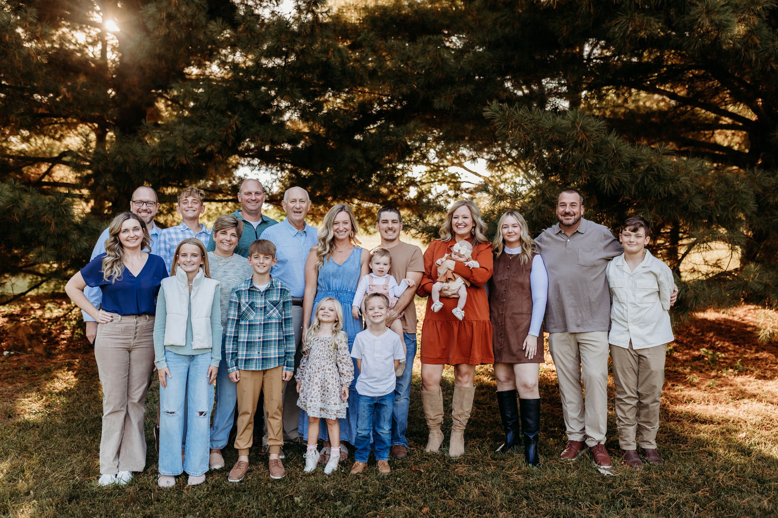 Large extended family group smiling together outdoors beneath tall pine trees in warm golden light.