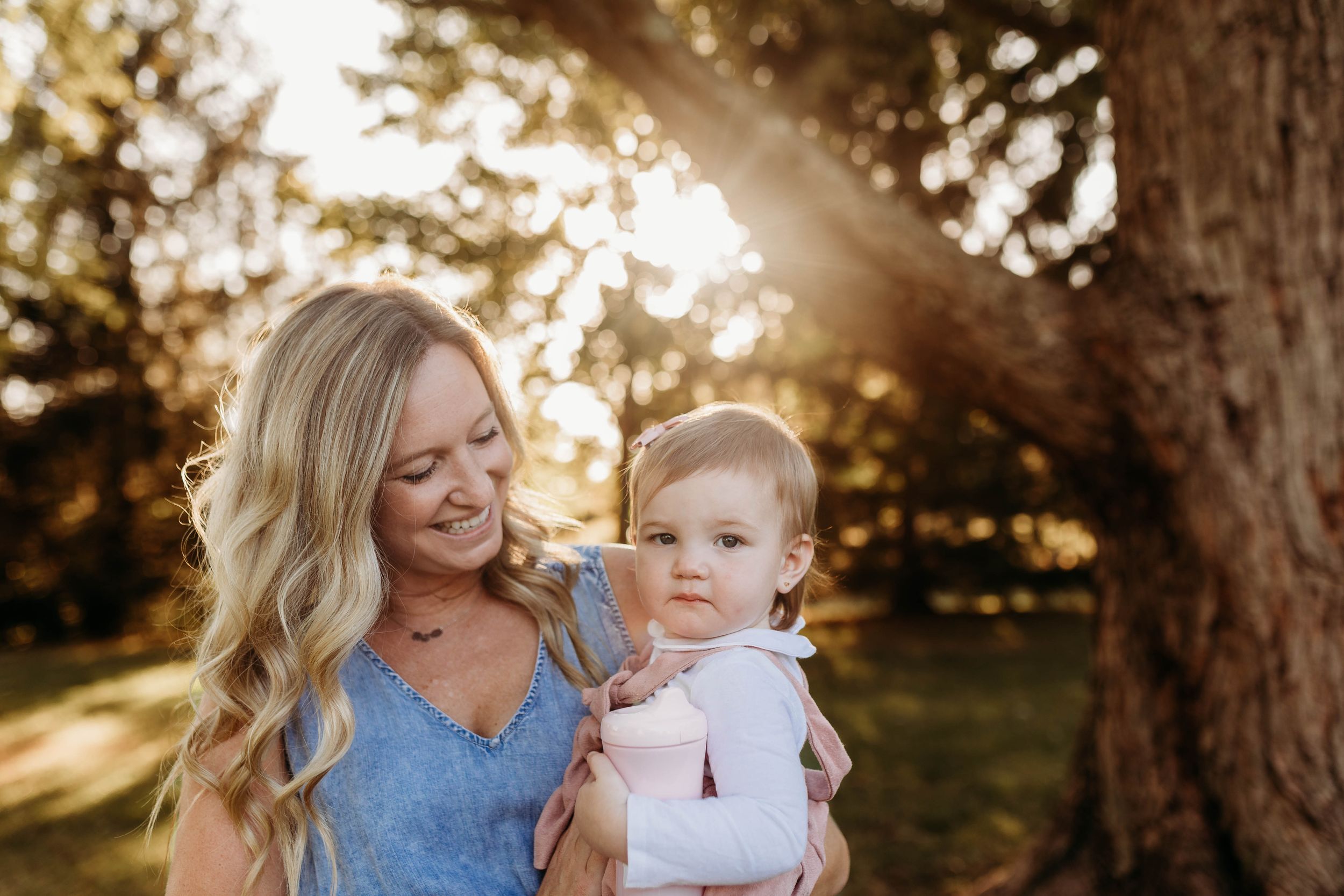 Smiling blonde mother holding adorable baby outdoors with sunlight filtering through trees behind them.