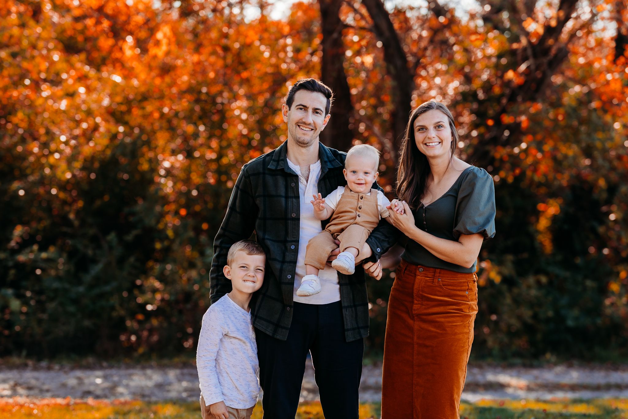 Smiling family of four stands together outdoors with colorful fall trees in background.