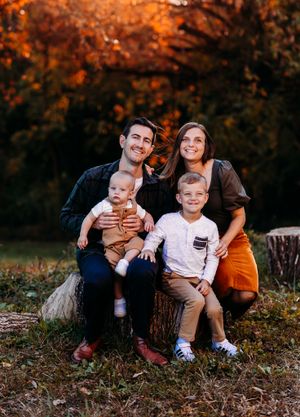 Family of four sits on a stump smiling together surrounded by golden autumn leaves.