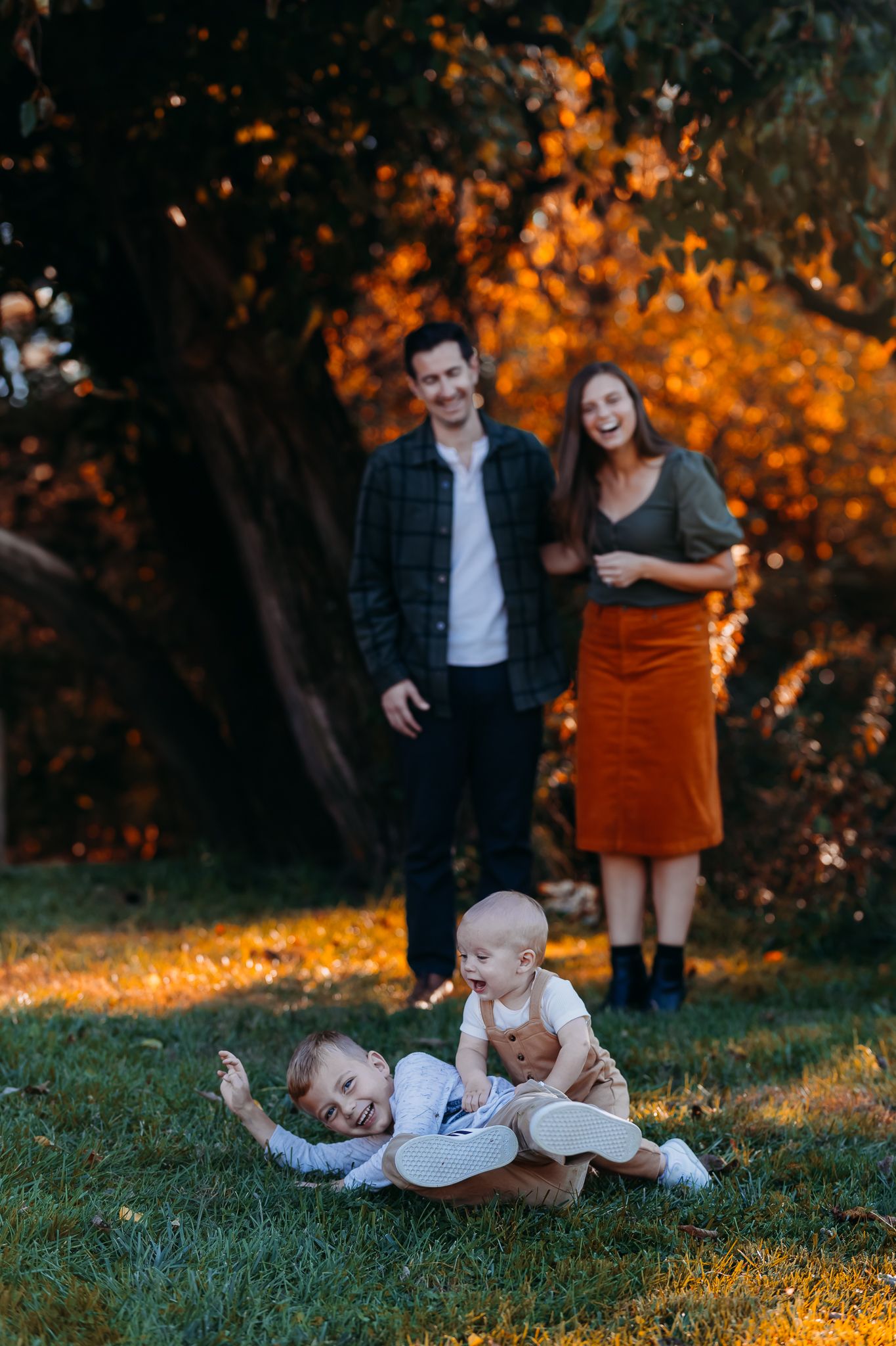 Two young siblings play on grass while parents laugh together in autumn setting.
