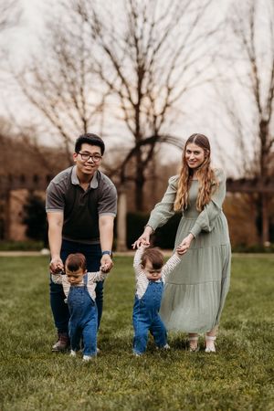 Parents in park helping twin toddlers in denim overalls take steps on grass.