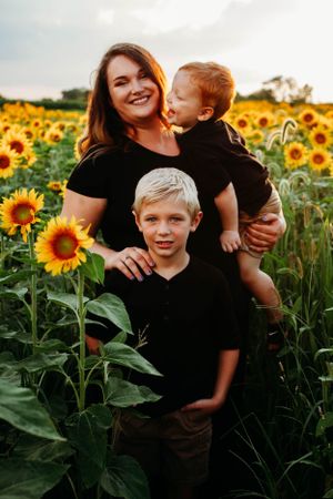 Smiling mother in black shirt holding toddler with older blonde boy in sunflower field at sunset.