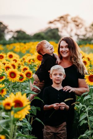 Happy mother in black dress holding laughing toddler with blonde boy in vibrant sunflower field.