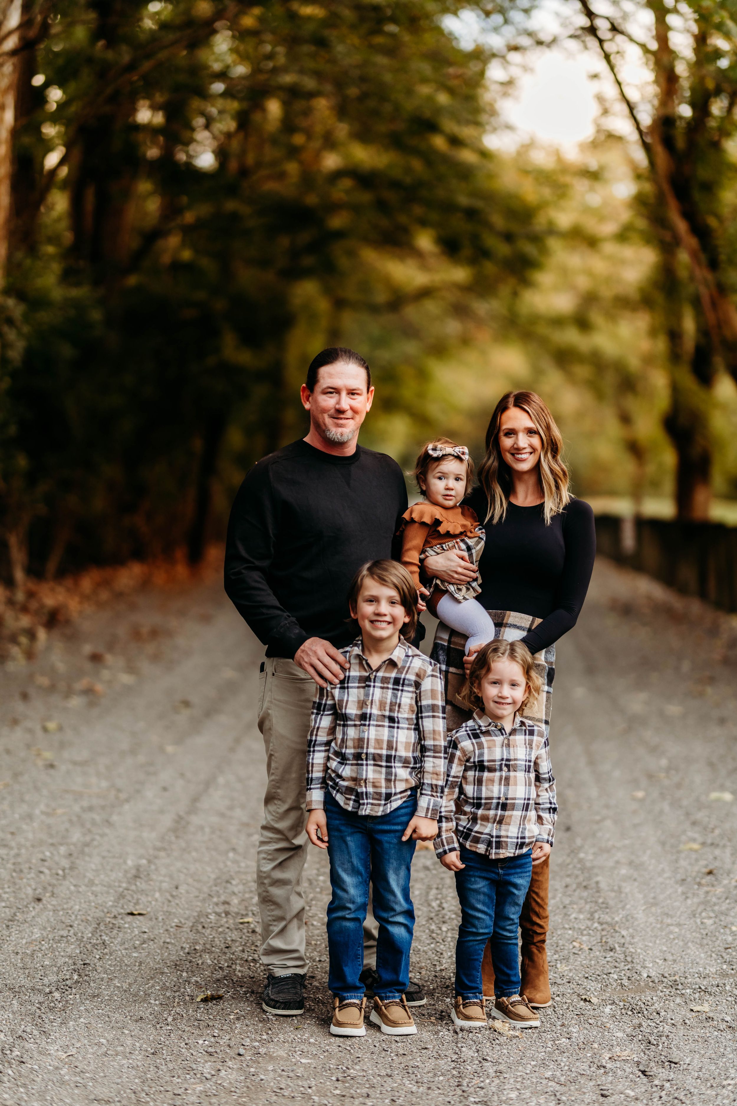 Family of five posing on a tree-lined gravel path during golden autumn sunset.