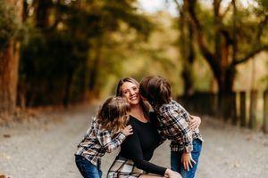 Mother crouching as two children in plaid shirts give her kisses on a gravel path.