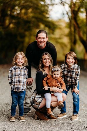 Family of five smiling together on a gravel path surrounded by warm autumn foliage.