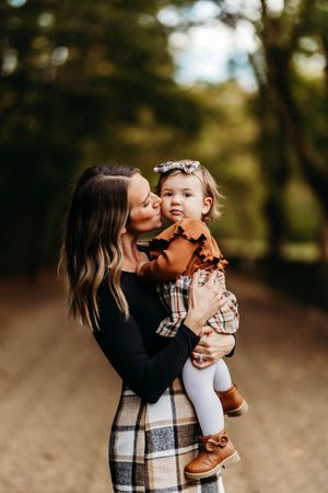 Mother kissing toddler daughter in rust orange outfit on a sunlit autumn path.
