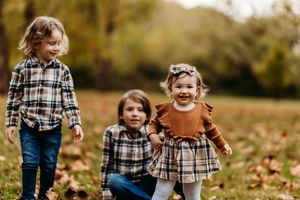 Three children pose outdoors in autumn, wearing coordinating plaid outfits among fallen leaves.