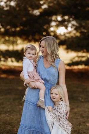 Blonde mother in blue dress holding toddler while young daughter hugs her side outdoors.