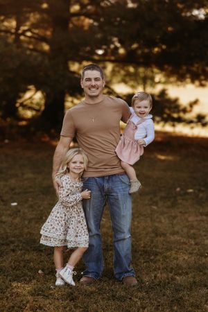 Smiling father in tan shirt holding baby girl while young blonde daughter stands beside him.