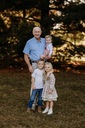 Elderly grandfather smiling outdoors holding baby while two young grandchildren stand in front.