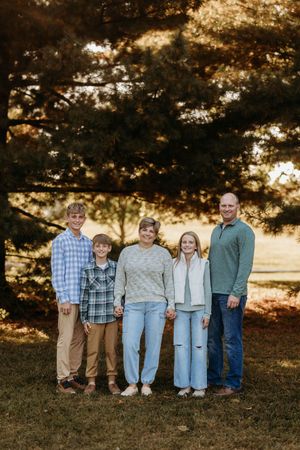 Family of five posing together outdoors under pine trees during fall golden hour photo session.