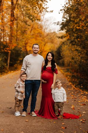 Family of four with pregnant mother in red gown posing on autumn leaf-covered path.