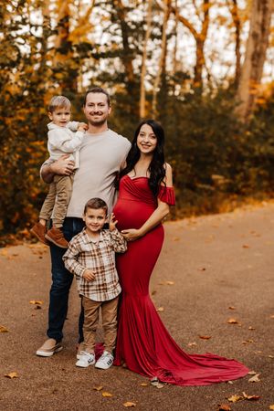 Expecting family with two young boys and pregnant mother in red maternity gown in fall foliage.