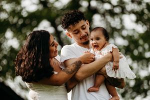 Young couple smiling and holding their happy baby girl outdoors with green trees in background.