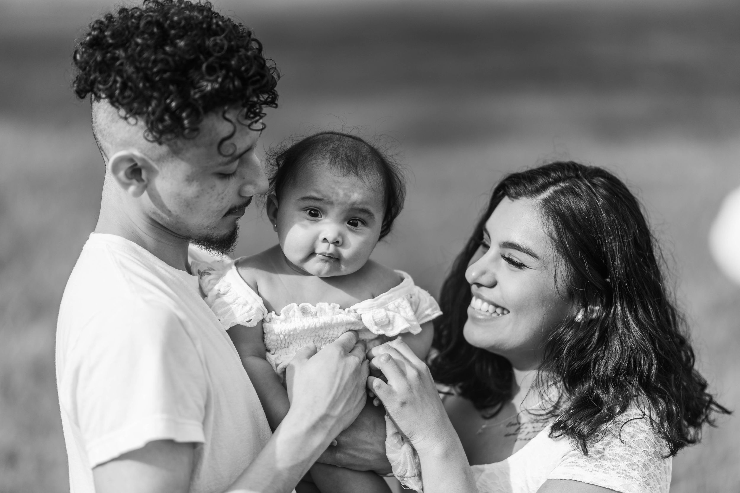 Black and white portrait of young parents lovingly holding their infant baby outdoors.