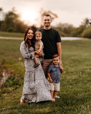 Smiling family of four stands in green field at golden hour near a pond.