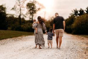 Family of four walking away on a gravel path at sunset, mother carrying baby, father holding toddlers hand.