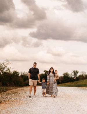 Happy family of four walking on a gravel path under dramatic cloudy sky, mother carrying baby while pregnant.