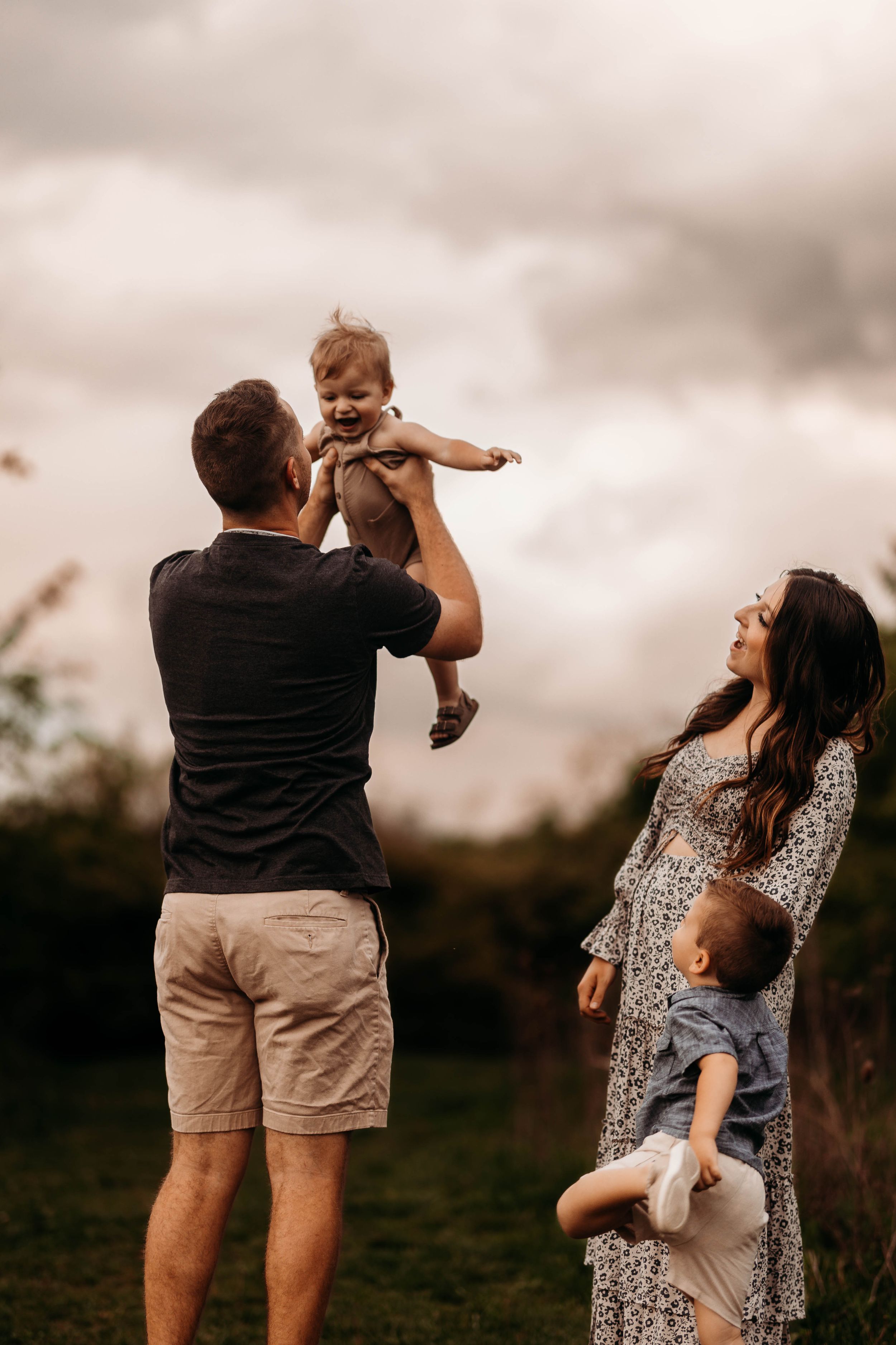 Father lifting laughing baby in the air outdoors while mother holds toddler against cloudy sky.