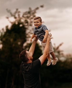 Joyful father tossing smiling toddler boy high in the air outdoors against a cloudy sky.