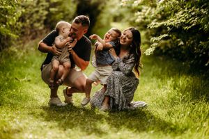 Happy family of four sitting on green grass path surrounded by lush trees, parents cuddling two young children.