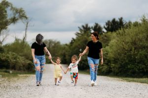 Two moms holding hands with two young children walking on a gravel path outdoors.