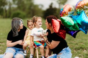 Two moms sitting on grass with two daughters and colorful birthday balloons.