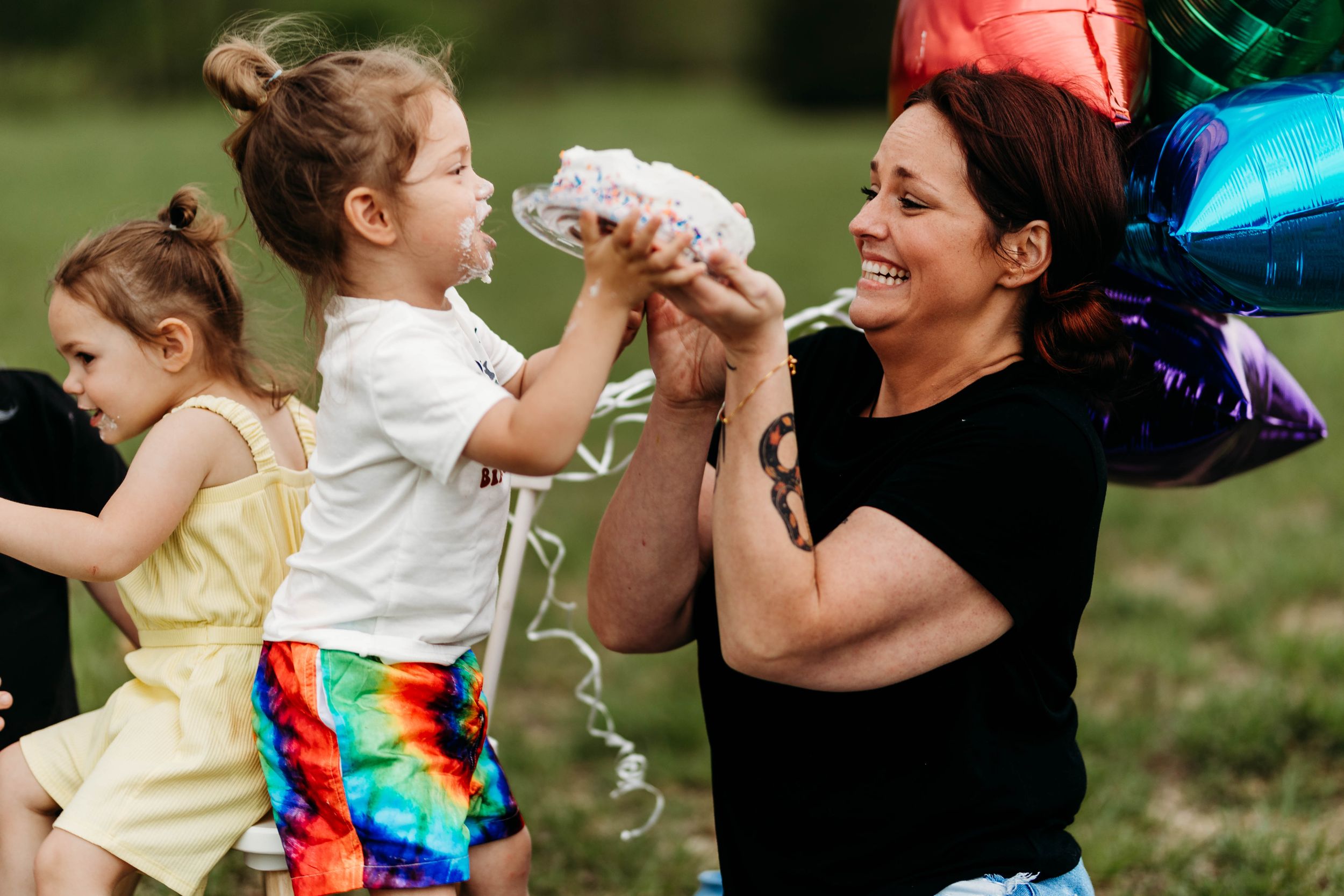 Mother laughing as young daughter hands her confetti during outdoor birthday celebration.