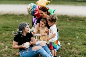 Family of two moms and two daughters celebrating with cake and balloons on grass.