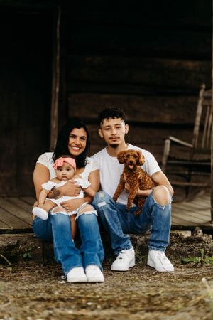 Young family sitting on rustic cabin steps, holding baby girl and brown poodle puppy.