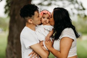 Young parents kissing their baby girl on the cheeks simultaneously outdoors near a tree.
