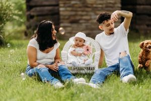 Young family with baby in wicker seat sitting on grass with dog, blowing bubbles outdoors.