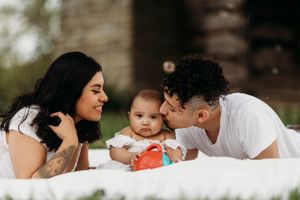Young couple lying on white blanket outdoors, lovingly looking at their smiling baby between them.
