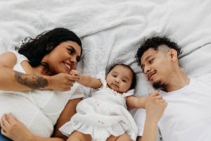 Happy family of three lying on white bed, parents smiling at adorable baby in white dress.