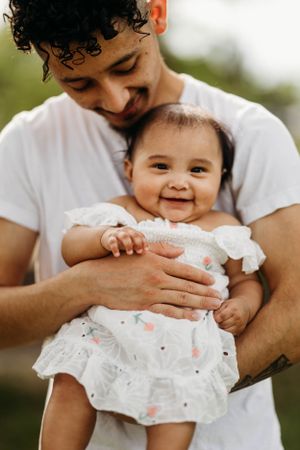 Smiling father in white t-shirt tenderly holding happy baby girl in white floral outfit outdoors.