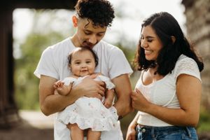 Smiling young family in white shirts, father holding baby girl in white dress outdoors.